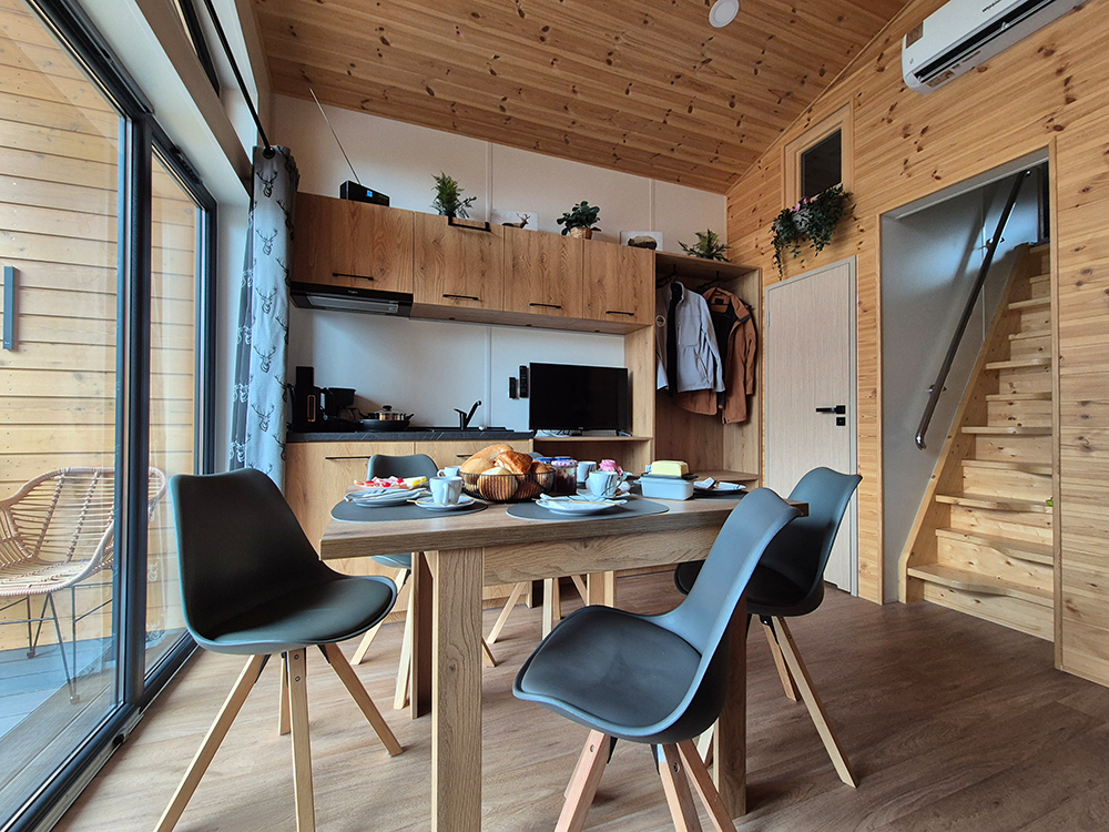 Moderner Essbereich im klimatisiertem Alpenchalet Wendelstein als kühler Rückzugsort im Sommer oder als warmes Nest nach im Winter mit offener Küche, Treppenaufgang und Blick durch die Panoramafront auf die hauseigene Terrasse mit freiem Blick auf den Wendelstein.
