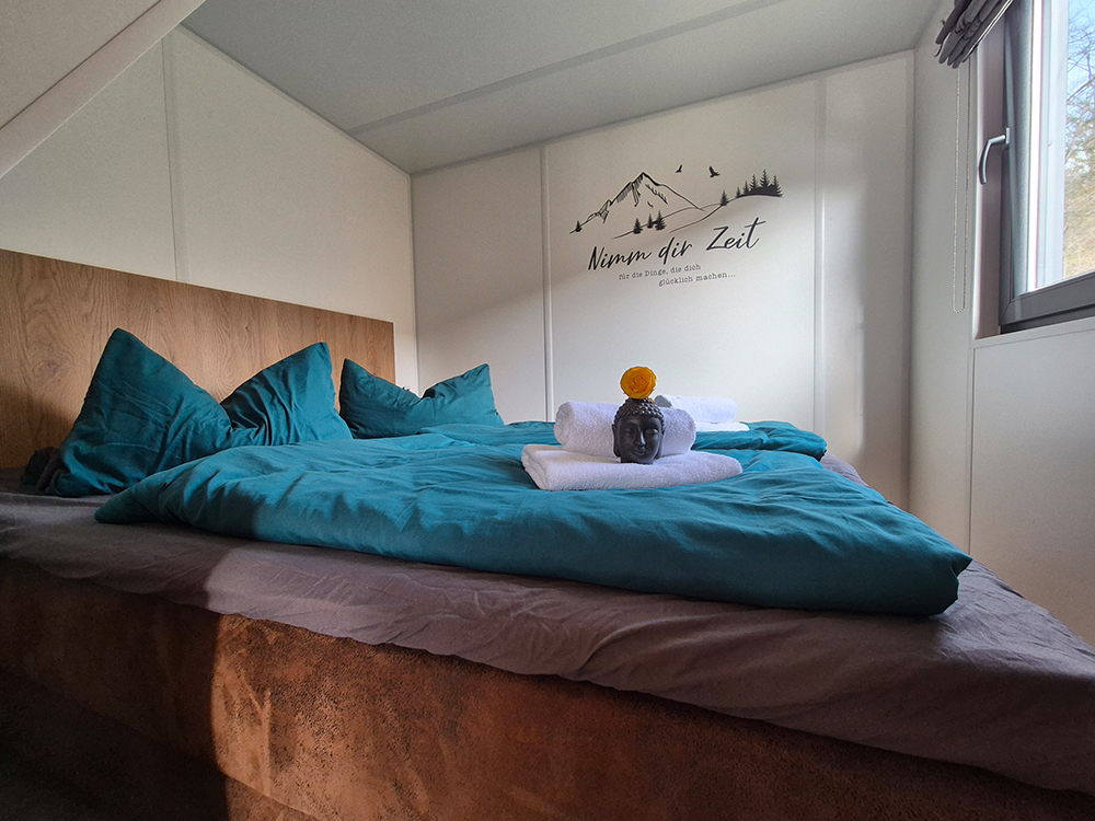 Ein sonnendurchflutetes Schlafzimmer im Alpenchalet Wendelstein mit Ostfenster und Morgensonne. Das Doppelbett ist mit petrolfarbener Bettwäsche bezogen. Auf den weißen Handtüchern in der Mitte ruht ein kleiner Buddha-Kopf mit einer gelben Blüte. An der Wand darüber befindet sich ein Wandtattoo mit Bergmotiv und dem Schriftzug „Nimm dir Zeit für die Dinge, die dich glücklich machen…“.