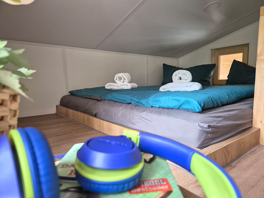 Ein gemütlicher Schlafplatz für zwei Personen im Obergeschoss des Alpenchalets Wendelstein. Das Doppelbett in einer hellen Holznische ist mit blaugrüner Bettwäsche bezogen; darauf liegen zwei kunstvoll gerollte weiße Handtücher. Im unscharfen Vordergrund sind ein Buch und blau-grüne Kopfhörer zu sehen, im Hintergrund ein kleines Holzfenster im charmant bayrischen Stil.